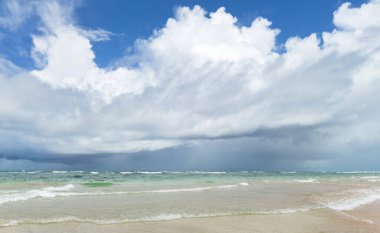Sakin suların kuma değdiği kıyı manzarası beyaz ve gri bulutlarla işaretlenmiş mavi gökyüzünün altında dramatik bir kontrastlık yaratıyor..