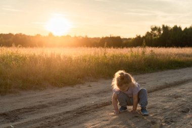 Küçük bir kız kumlu toprak bir yolda oynuyor. Yazın gün batımında kumlu bir arazide.