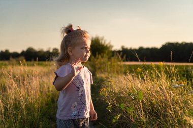 Küçük bir kız ilginç bir şekilde tarlanın ortasında duruyor ve uzaklığa bakıyor. Güneş bir yaz akşamında yüzünü kolayca aydınlatıyor.