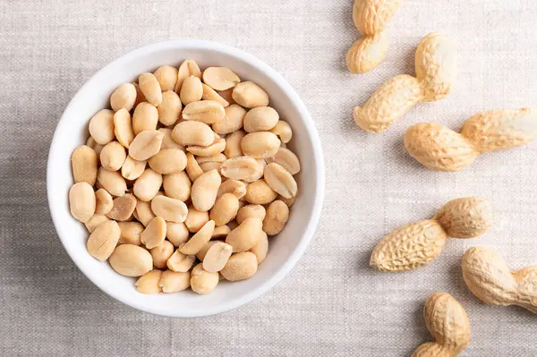 Stock image Roasted and salted peanuts in a white bowl on linen fabric. Ready-to-eat snack food, made from fruits of Arachis hypogaea, also known as groundnut, goober, pindar or monkey nut. Close-up from above.