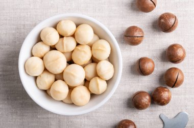 Beyaz bir kasede keten kumaşın üzerinde kavrulmuş macadamia fındığı. Doğru meyveler, doğranmış fındık kabukları ve anahtar. Queensland, Bush, Maroochi, Bauple ve Hawaii fıstıkları olarak da bilinir. Yakından, yukarıdan..