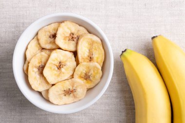 Muz cipsi ya da beyaz bir kasede keten kumaş üzerinde muz cipsi. Sarı çıtır çıtır, bol yağda kızartılmış muz dilimleri, şeker ya da bal ile kaplı. Tatlı bir atıştırmalık. Yakından, yukarıdan, yemek fotoğrafı..