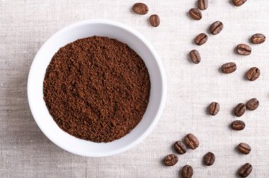 Kahve tozu, keten kumaşın üzerinde beyaz bir kasede taze kahve çekirdekleri. Sağdaki kavrulmuş çilek tohumlarında da Arap, dağ ya da Arabika kahvesi var. Yakından, yukarıdan..