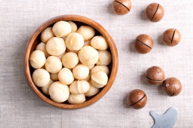 Roasted macadamia nuts in a wooden bowl on linen fabric. On the right fruits with sawn nutshells and opener key. Also known as Queensland, bush, maroochi, bauple and Hawaii nuts. Close-up, from above. clipart