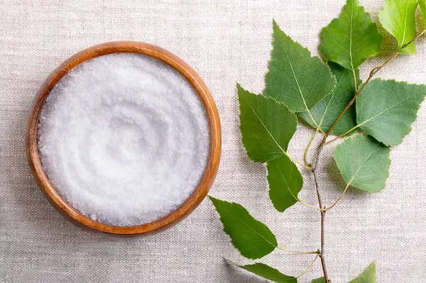 stock image Birch sugar, table sweetener based on xylitol, in a wooden bowl on linen fabric. Also called xylite, a sugar substitute of natural origin with 40 percent fewer calories. A birch branch on the left.