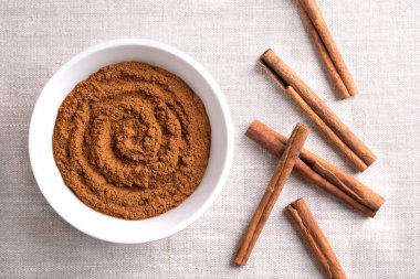 Cinnamon powder, in a white bowl on linen fabric. Raw bark of Indonesian cinnamon, Cinnamomum burmannii, ground and whole. Used mainly as aromatic condiment and flavouring additive in a wide variety. clipart