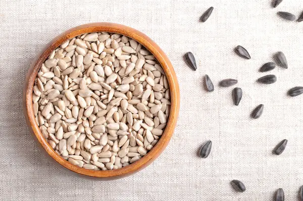 stock image Hulled sunflower seeds in a wooden bowl on linen fabric. Fruits of the oilseed Helianthus Annuus, the common sunflower. On the right side whole seeds with black hulls. Close-up from above, food photo.