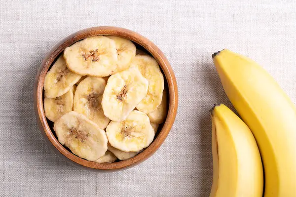 stock image Banana chips or banana crisps in a wooden bowl on linen fabric. Yellow crunchy deep fried slices of bananas, covered with sugar or honey. Snack with sweet taste. Close-up, from above, food photo.