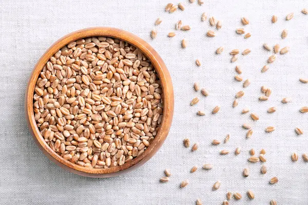 stock image Common wheat kernels, bread wheat in a wooden bowl on linen fabric. Triticum aestivum, a grass widely cultivated for its seed, a cereal grain and a staple food around the world. Close up from above.