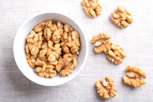 Stock image Walnut kernel halves in a white bowl on linen fabric. Shelled and dried halves of walnut kernels, ripe seeds of common walnut tree Juglans regia, used as snack or for baking. Close-up from above.