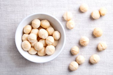 Macadamia nuts, roasted and salted, in a white bowl on linen fabric. Also known as Queensland nut, bush nut, maroochi nut, bauple and Hawaii nuts. Ready-to-eat snack nuts. Close-up, from above. Photo clipart