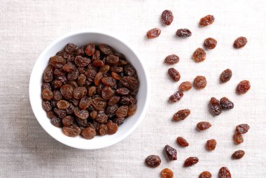 Genel ticari kuru üzüm, keten kumaş üzerine beyaz bir kase içinde. Koyu renkli büyük çekirdeksiz üzümler, çiğ yenmek ya da pişirmek, pişirmek ve demlemek için kullanılır. Yakından, yukarıdan. Yemek fotoğrafı..