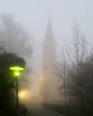 Yeşil fenerli kalın sis altındaki kilise. Guggenthal Şube Kilisesi, Kasım akşamının sisinde. Avusturya 'nın Koppl kentindeki Salzburg belediyesinde Kutsal Haç' a adanmış Roma Katolik Kilisesi.
