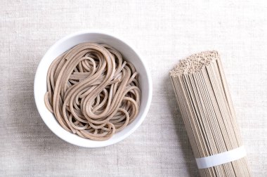 Soba eriştesi, kaynatılmış ve soğutulmuş, keten kumaşın üzerinde beyaz kâsede. İnce Japon eriştesi, başlıca karabuğday unundan yapılmış, buğday unuyla karıştırılmış. Sağında kurutulmuş erişte paketiyle..