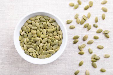 Pepitas, kabuklu balkabağı tohumları, beyaz bir kasede, keten kumaşın üzerinde. Çiğ, kurutulmuş kabak tohumu, Cucurbita meyveleri kabuk çıkarıldıktan sonra açık yeşil ve aperatif, garnitür ya da malzeme olarak kullanılır. Fotoğraf.