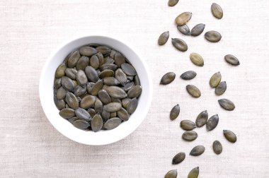 Beyaz bir kase içinde keten kumaş üzerinde, yapay yağ kabağı tohumları. Kurumuş, koyu yeşil ve taze meyveli balkabağı, Cucurbita peposu. Styriaca, tohum yağı için yetiştirilmiş sıradan bir balkabağı çeşidi..