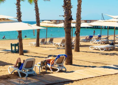 Antalya; 10 Eylül 2022: Güneşli yataklarda dinlenen ve plajdaki palmiye ağaçlarının altında güneşlenen turist çift. Arka planda denizde yüzen insanlar. Antalya Rus, Ukraynalı, İngiliz, Polonyalı ve Alman turistler için popüler bir şehirdir. 