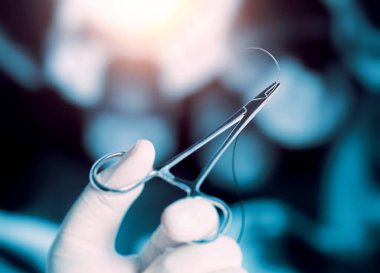 Operatör yarım daire şeklinde dikiş ve iğne kesiyor. Hemostatik forseps ile ameliyat odasındaki ışıkların altındaki bulanık operatörlerin önünde..