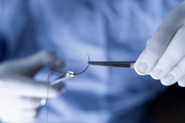 Dikişlerle ilgilenen mavi önlüklü bir tıp uzmanı. Operatör operasyon sırasında pens ve iğne tutacağı ile yarım daire iğnesi tutuyor.