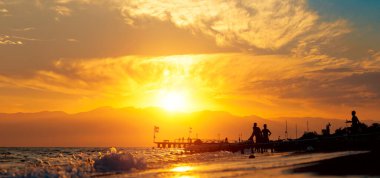 Panoramic summer background with silhouettes of people by the sea.  Beach-goers at the seashore during golden hour in summer in Antalya, Turkey.
