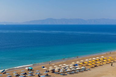 İnsanlar Yunanistan 'ın Rhodes Adası' ndaki Elli Sahili 'nde yüzüyor, güneşleniyor veya başka aktivitelerde bulunuyorlar. Kumda renkli şemsiyeler var. Ufuktaki dağlar.