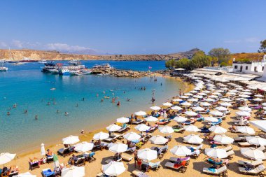 LINDOS-GREECE: 26 Ağustos 2024: Yunanistan 'ın Rodos kentindeki Lindos Plajı, altın kum, turkuaz sular, güneşlik yatakları ve resimli bir koyda demirlemiş teknelerle övünüyor.