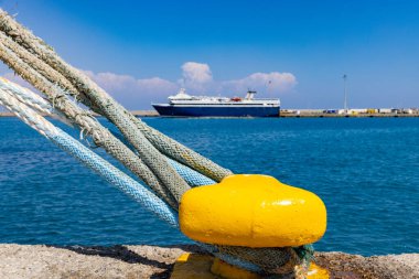Parlak mavi bir gökyüzüne ve sakin bir denize karşı yanaşmış bir gemiyi koruyan kalın halatlarla bağlanmış sarı bir korkuluğun yer aldığı canlı bir liman sahnesi. Seyahat konsepti.