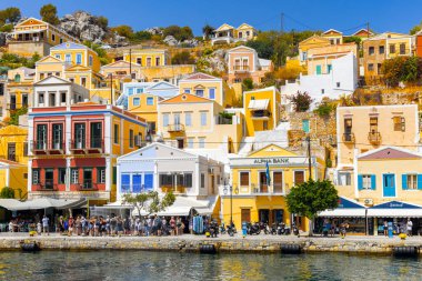SYMI-GREECE: 27 Ağustos 2024: Ege Denizi 'nin mavi sularına bakan renkli neoklasik evler Yunanistan' ın Symi Adası 'nın yamacında yer alıyor. Turistler rıhtım boyunca toplanarak canlı manzaranın ve yerel dükkanların tadını çıkarıyorlar..