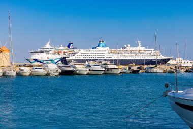 RHODES-GREECE: 25 Ağustos 2024: Yunanistan 'ın Rodos Adası' ndaki tarihi Mandraki Limanı 'na yanaşan görkemli yolcu gemileri, pırıl pırıl gök mavisi sular ve canlı bir sahil atmosferiyle çevriliydi..