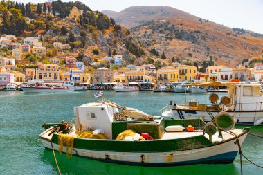 Symi Adası 'ndaki liman sahnesi, renkli tekneler yanaştı. Canlı binaları olan tepe kenarı kasabası denize bakıyor..