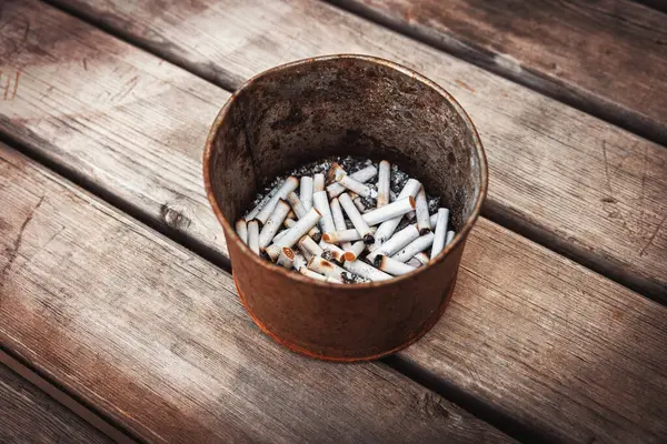 Sayısız sigara izmariti ve külle dolu metal bir kap yıpranmış ahşap bir yüzeye oturur. Sağlıksız bir alışkanlığın ardından ortaya çıkan sert bir fotoğraf..