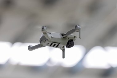 Bir kompakt, 249 gramlık gri İHA alçaktan uçuyor, pervaneleri hafifçe bulanıklaşıyor, bir iç uzay içinde aktif olarak video kaydettiğini gösteriyor. Odak, İHA 'nın parlak tasarımını ve hava görüntüleri için ima edilen işlevini vurgular.