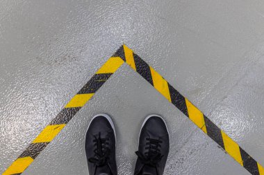 Siyah ayakkabılı bir insanın ayakları, belirgin sarı ve siyah çizgili tehlike uyarısı çizgisi ile çevrili gri beton bir zeminde durur. Bu görüntü güvenlik protokollerini ve fabrika ortamında belirlenmiş alanları gösterir.