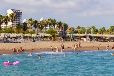 28 Temmuz 2023 'te insanlar kalabalık bir plajda palmiye ağaçları ve büyük bir otel kompleksi olan güneşli bir günün tadını çıkarıyorlar. Tatilciler güneşlenirken, voleybol oynarken ve Antalya 'da sakin denizde yüzerken görülüyor..