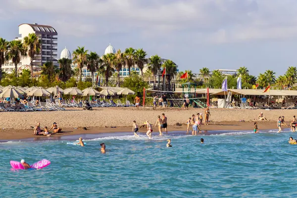 28 Temmuz 2023 'te insanlar kalabalık bir plajda palmiye ağaçları ve büyük bir otel kompleksi olan güneşli bir günün tadını çıkarıyorlar. Tatilciler güneşlenirken, voleybol oynarken ve Antalya 'da sakin denizde yüzerken görülüyor..