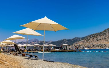 Selimiye-Marmaris: 30 Ağustos 2024: Huzurlu bir sahildeki büyük şemsiyeli şezlonglar, suyun üzerindeki ahşap iskeleye uzanıyor. Berrak, sakin körfez dağlarla çevrili, uzakta bir tekne var..