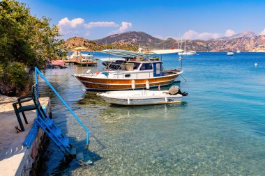 Selimiye-Marmaris: 30 Ağustos 2024: Kristal berraklığında bir körfezde mavi korkuluklu bir taş iskelenin yanında iki tekne demirli. Sakin sahne, uzak tepeler ve parlak mavi gökyüzü tarafından tamamlandı..