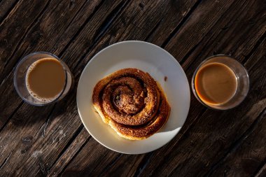 A sticky bun in the sunlight on a wooden table and two cups of coffee.