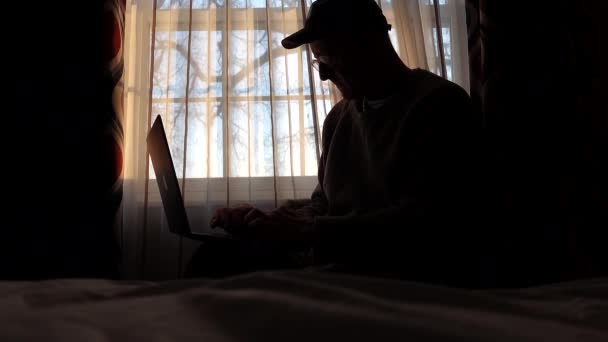一名中年男子 头戴帽子 在宾馆房间里 黎明时分在窗下的电脑上工作 — 图库视频影像