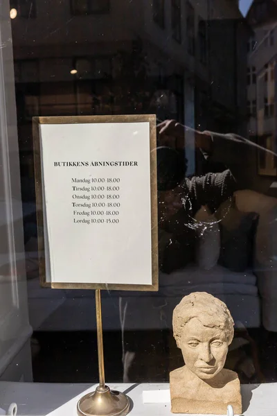 Copenhagen, Denmark A  storefront window sign in the old town announces opening hours and a samall stone head statue.