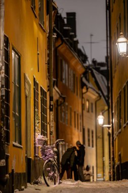 Stockholm, İsveç 28 Kasım 2023. Old Town veya Gamla Stan 'in dar karlı sokaklarında geceleri yürüyen insanlar ve park halindeki bir bisiklet..