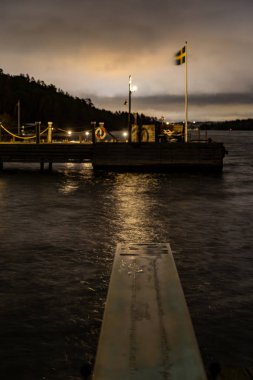 Stockholm, İsveç Ekero banliyösünde geceleri Malaren Gölü 'nde küçük bir rıhtım rüzgarda dalgalanan bir İsveç bayrağı..