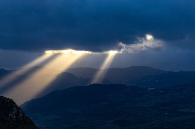 Caccamo, Sicilya, İtalya Gün batımında dağları aydınlatan bulutların arasından güneş ışığı parlıyor.