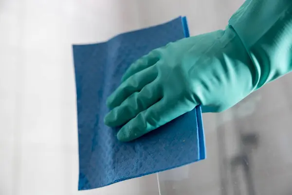 A hand in a rubber gloves cleans a shower stall and glass cabin.