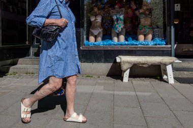 Stockholm, İsveç 21 Mayıs 2024. Mariatorget 'te, yazlık elbiseli yaşlı bir kadın bikini mağazasının önünden geçiyor..