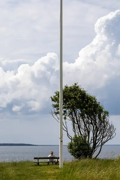 Stock image Hundested, Denmark May 27, 2024 A woman sits on a bench overlooking the Baltic Sea and Kattegat straits on a cloudy day under a flagpole.