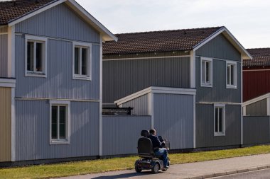 Stockholm, Sweden A man in an elecctric wheelchair rolls by houses on Prastgardsvagen in the Tumba suburb. clipart