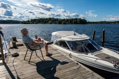 Stockholm, Sweden A man sits on a small boat pier in Lake Malaren next to a motorboat. clipart