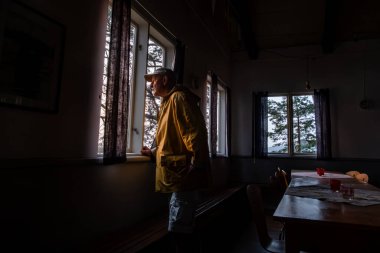 Stockholm, İsveç. Sarı yağmurluklu bir adam şafak vakti gölün kenarındaki küçük bir kulübede pencereden dışarı bakar..