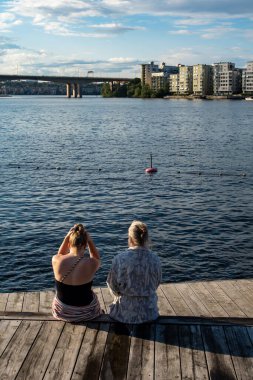 Stockholm, İsveç 31 Temmuz 2024. İki kadın gün batımında, şehir merkezi ve Malaren Gölü manzaralı ahşap bir güvertede oturur..